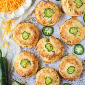 Cheddar jalapeño biscuits spread out on a white surface with a plate of cheddar cheese in the top left corner.