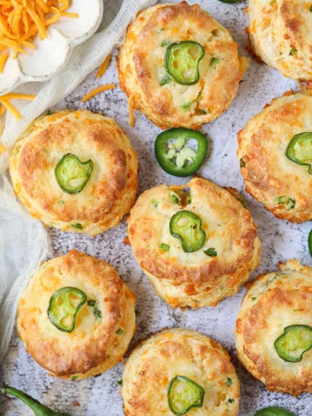 Cheddar jalapeño biscuits spread out on a white surface with a plate of cheddar cheese in the top left corner.
