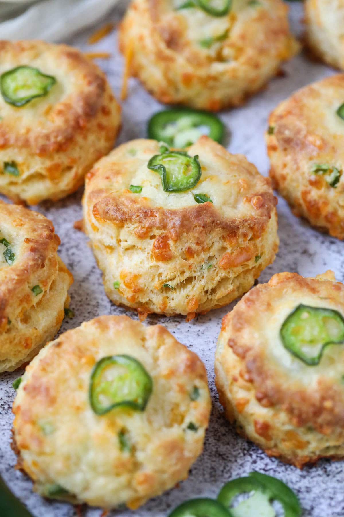 Cheddar jalapeño biscuits spread out on a white surface with a plate of cheddar cheese in the top left corner.