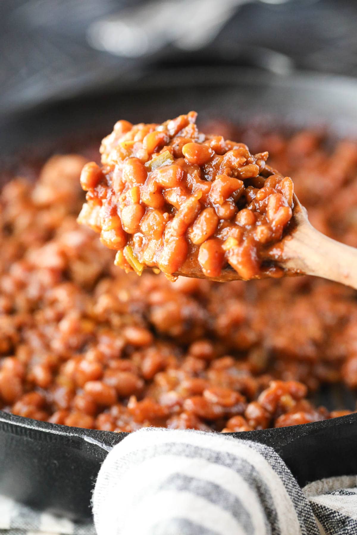 A spoonful of Southern baked beans being held over a cast-iron skillet.