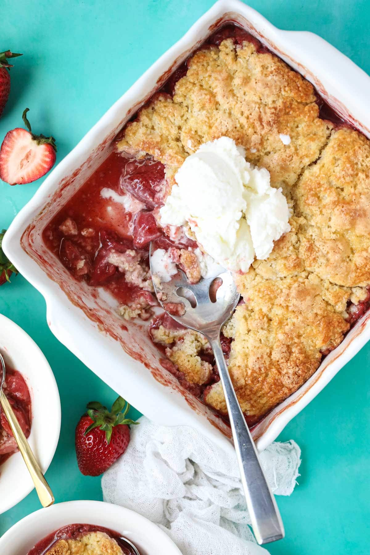 Fresh strawberry cobbler in a white baking dish sitting on a blue surface.