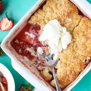 Fresh strawberry cobbler in a white baking dish sitting on a blue surface.