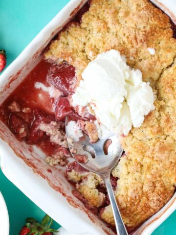 Fresh strawberry cobbler in a white baking dish sitting on a blue surface.