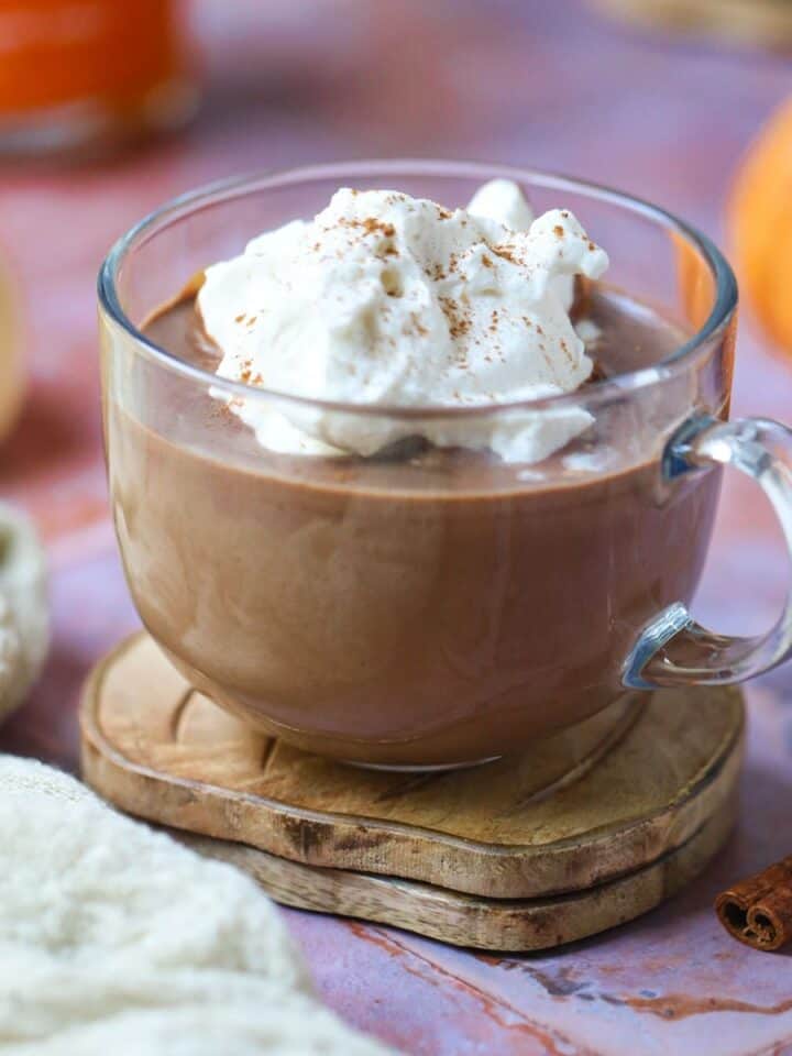 Pumpkin hot chocolate in a clear mug topped with whipped cream sitting on a copper tile surface.