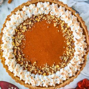 Pumpkin tart topped with toasted meringue and chopped walnuts sitting on a white cake plate on top of a gray marble surface.