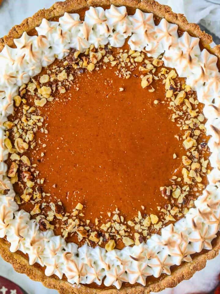 Pumpkin tart topped with toasted meringue and chopped walnuts sitting on a white cake plate on top of a gray marble surface.