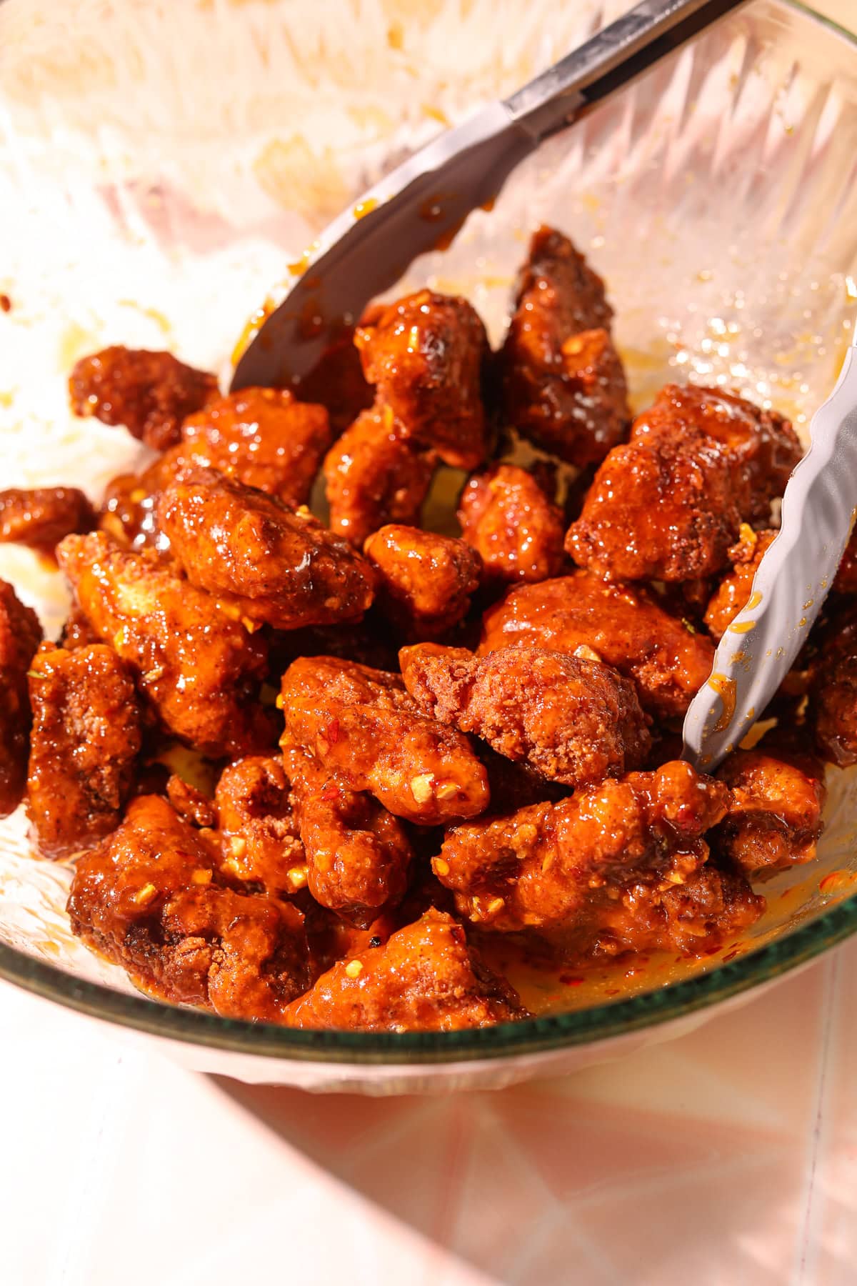 Tossing breaded chicken bites in hot honey garlic sauce in a bowl with tongs.